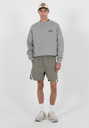 Giovane uomo con felpa grigia, pantaloncini sportivi verde oliva, calzini bianchi, scarpe da ginnastica e cappellino beige, in piedi con le mani nelle tasche.