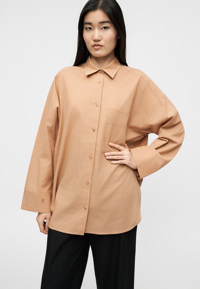 Γυναίκα με μακριά σκουρόχρωμα μαλλιά, ντυμένη με υπερμεγέθη μπεζ πουκάμισο με κουμπιά και μαύρο παντελόνι, στέκεται με το ένα χέρι στη μέση.
