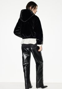 Černá krátká bunda z umělého kožíšku s kapucí, doplněná lesklými černými koženými kalhotami. Silueta je volná a zakončená shrnutým lemem.