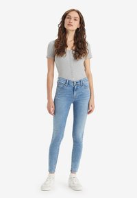 Γυναίκα με μακριά σγουρά μαλλιά, φορώντας γκρι κοντομάνικο πουκάμισο με κουμπιά, ανοιχτό μπλε στενά τζιν, λευκές κάλτσες και λευκά αθλητικά παπούτσια, όρθια.