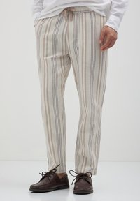 Homme portant un pantalon rayé vertical beige et marron avec une taille à cordon et des chaussures bateau en cuir marron foncé à lacets.
