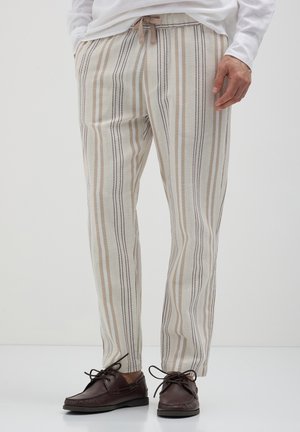 Uomo che indossa pantaloni a righe verticali beige e marroni con vita regolabile e scarpe da barca in pelle marrone scuro con lacci.