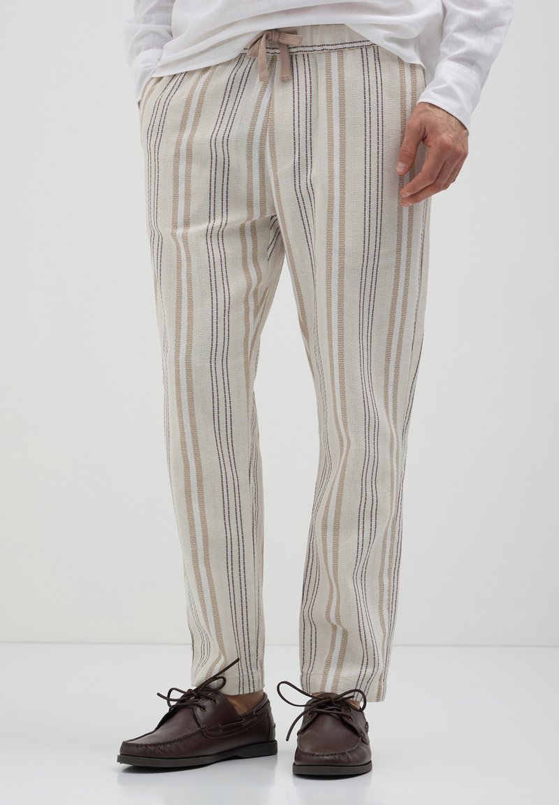 Homme portant un pantalon rayé vertical beige et marron avec une taille à cordon et des chaussures bateau en cuir marron foncé à lacets.