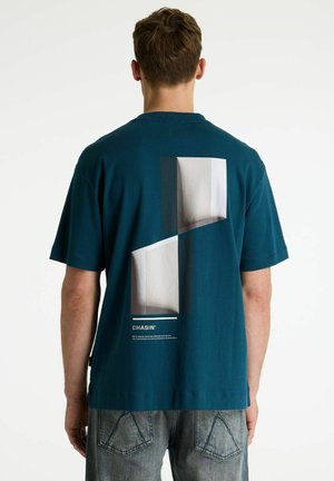 Homme portant un t-shirt bleu sarcelle avec un motif abstrait gris et blanc et le texte "CHASIN'" dans le dos, associé à un jean bleu délavé.