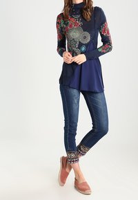 Marineblaues Langarmshirt mit floraler Stickerei auf den Ärmeln, kombiniert mit eng anliegenden Jeans; die Jeans haben einen dekorativen Saum. Lässige Schuhe vervollständigen den Look.