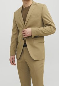 Beige kostym med en enfärgad jacka och slagkrage, två framfickor och matchande smala byxor. En strukturerad brun tröja under.