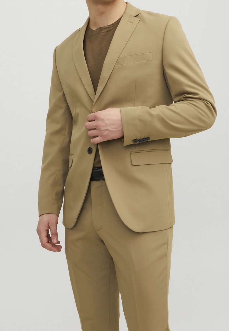 Beige kostym med en enfärgad jacka och slagkrage, två framfickor och matchande smala byxor. En strukturerad brun tröja under.