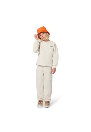 Enfant portant un sweatshirt matelassé crème et un pantalon assorti, un chapeau seau orange et des chaussures beiges avec des motifs de visage de tigre, debout et touchant son chapeau.