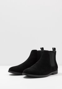 Stivali alla caviglia in suede nero con pannelli elastici sui lati e linguette per il sollevamento. Suola piatta con una finitura liscia, caratterizzata da un design con punta arrotondata.