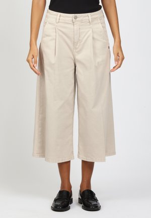 Woman wearing beige wide-leg cropped pants and black loafers, standing with arms relaxed at sides against a plain background.