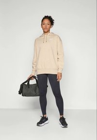 Beige hoodie med dragsko, logotyp på bröstet, matchad med svarta leggings och svarta sneakers. Håller en mörkgrön duffelväska.