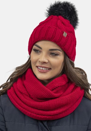 Rød strikket hat med en sort pom-pom, parret med et kraftigt rødt tørklæde, begge med tykke, strukturerede mønstre. Varme og hyggelige materialer.