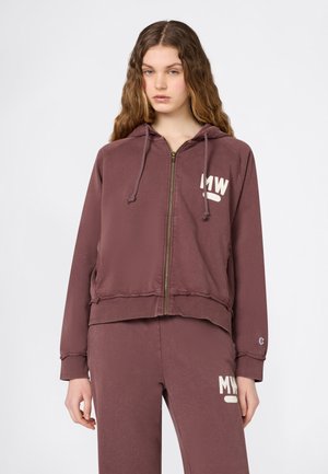 Giovane donna con capelli lunghi e mossi che indossa una felpa con zip e pantaloni coordinati in mauve polveroso con loghi "MW" sul petto e sulla coscia.