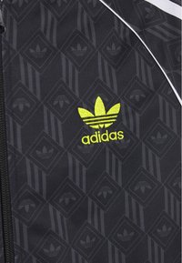 Chaqueta negra con un patrón de diamantes que presenta logotipos de Adidas. El material tiene una textura suave. Costuras en amarillo neón acentúan el diseño.
