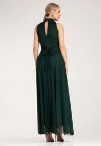 Długa, ciemnozielona sukienka z dopasowanym górnym partem i zwiewnym dołem. Posiada wysoki kołnierz, odkryte plecy oraz detal w postaci wiązania w talii.