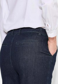 Jeans en denim bleu foncé présentant une coupe slim, des coutures discrètes et des poches latérales. Ils ont une taille mi-haute et une texture lisse.