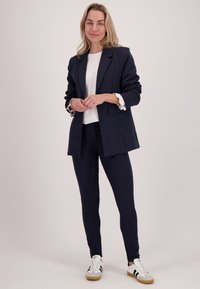 Vrouw standing in een marineblauw gestreepte blazer, witte top, marineblauwe broek en witte sneakers tegen een effen lichte achtergrond, met de handen losjes samengevouwen.