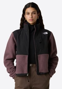 Giacca in pile nera e viola scuro con colletto alto, zip frontale e tasche laterali. Presenta un piccolo logo sulla spalla e maniche testurizzate.