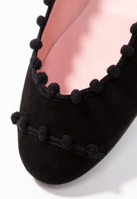 Chaussure en daim noir avec un bout rond, dotée d'un bord festonné et de petits accents de pompon noir le long du bord. Doublure intérieure rose.