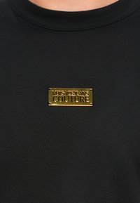 Μαύρο πουκάμισο με μια μικρή ορθογώνια χρυσή μεταλλική ετικέτα κεντραρισμένη στο στήθος, χαραγμένη με "VERSACE JEANS COUTURE" σε κεφαλαία γράμματα.