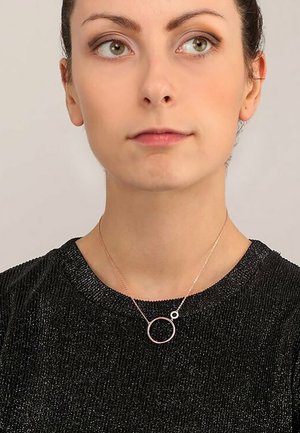 Roségoldene kreisförmige Halskette an einer dünnen Kette, getragen über einem schwarzen metallic-texturierten Oberteil. Sie weist einen größeren Kreis mit einem kleineren Akzentkreis auf.