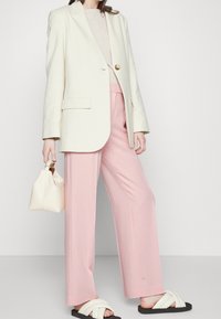 Krämfärgad kavaj med guldkknappar, ljusrosa vida byxor och svarta snörningsfria skor. Liten krämfärgad handväska med rund form.