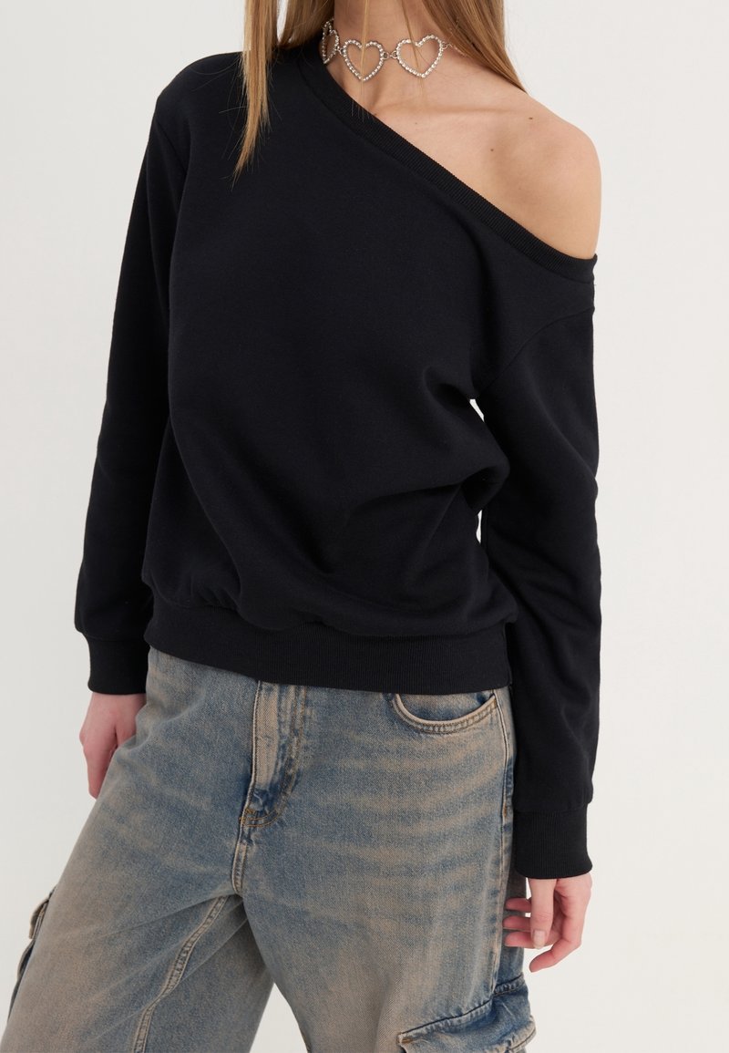 Femme portant un sweat-shirt noir à épaules dénudées, un pantalon cargo en denim bleu clair et un collier ras-de-cou argenté en forme de cœur sur fond uni.