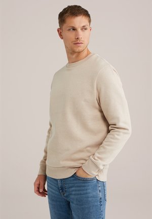Mlad moški gleda stran, oblečen v bež sweatshirt in modre jeans, z desno roko v žepu, nevtralen ozadje.