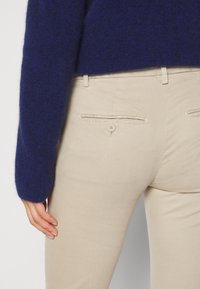 Beige Hosen mit Taschen auf der Rückseite und Knopfverschluss, kombiniert mit einem blauen Pullover. Der Stoff erscheint glatt und hat eine figurschmeichelnde Passform.