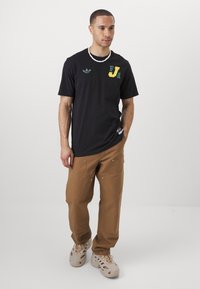 Black short-sleeve t-shirt featuring green and yellow accents, round neckline, and subtle logo on the chest. Worn with light brown pants.