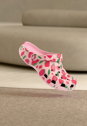 Sabot rose avec un motif fraise et cerise, des trous de ventilation et une bride au talon, positionné au-dessus d'un tapis beige dans une pièce neutre.