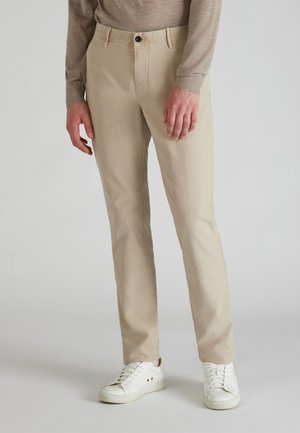 Beige Chinos aus Baumwolle mit geradem Schnitt, ausgestattet mit einem vorderen Knopfverschluss, Seitentaschen und getragen mit weißen Sneakers.