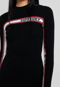 Maglione nero a coste con maniche lunghe, caratterizzato da un logo centrale "SUPERDRY" e strisce rosse e bianche in contrasto lungo i lati.