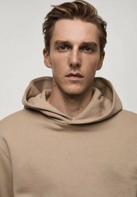 Bež hoodie s vezicom, s mekanom teksturom i opuštenim krojem. Kapuljača je istaknuta, što nadopunjuje minimalistički dizajn.