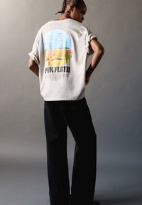 Osoba z krótkimi warkoczami, ubrana w biały T-shirt z grafiką zespołu Pink Floyd i czarne spodnie, na jednolitym szarym tle.