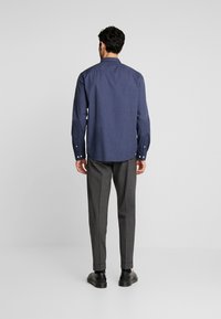 Camicia blu navy con bottoni, a maniche lunghe e un motivo testurizzato, abbinata a pantaloni grigi su misura e scarpe nere.