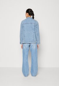 Woman with dark braided hair wearing light blue denim jacket and wide-leg jeans, standing facing a plain white wall.