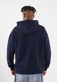 Persona con dreadlocks corti, orecchini a cerchio, che indossa una felpa blu navy e jeans di denim blu chiaro, girata di spalle su uno sfondo bianco.