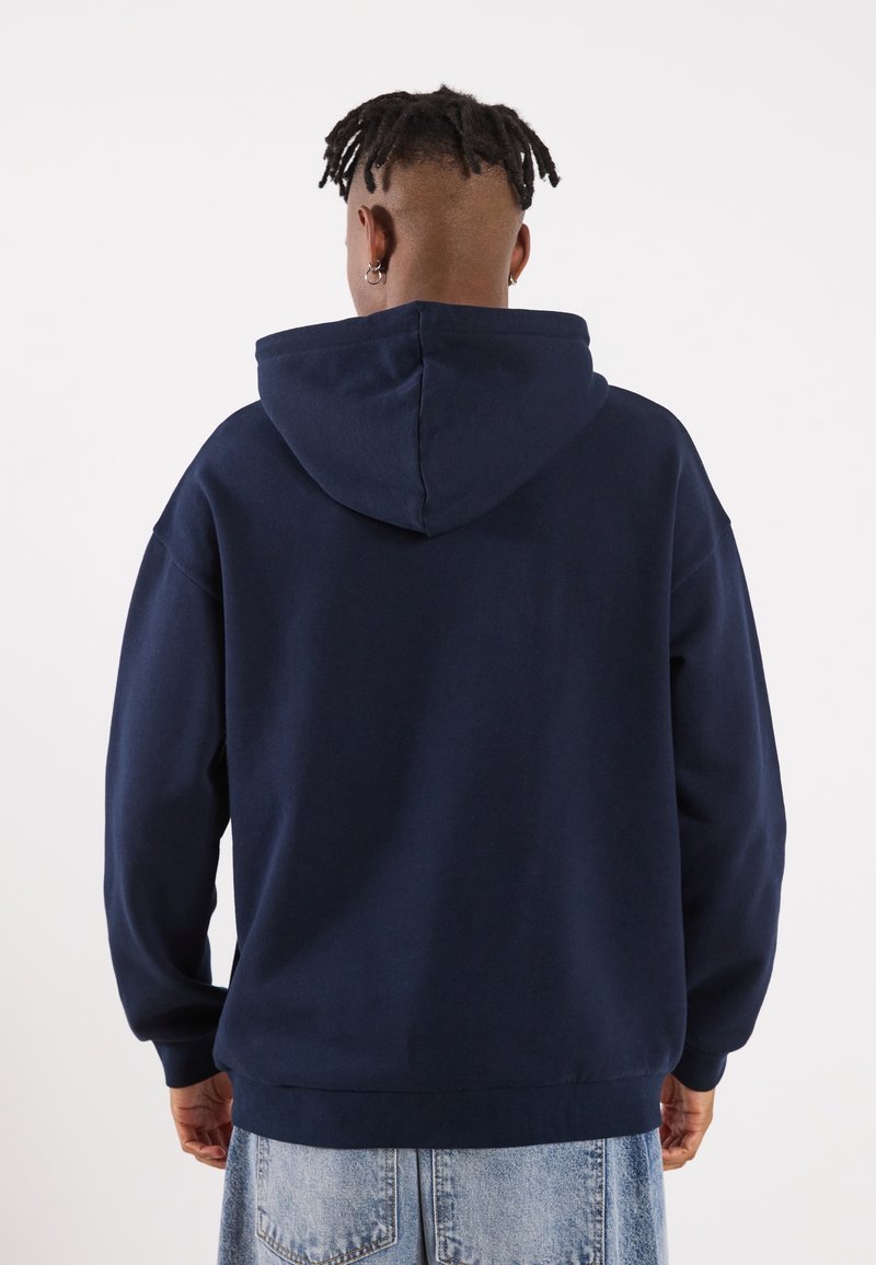 Persona con dreadlocks corti, orecchini a cerchio, che indossa una felpa blu navy e jeans di denim blu chiaro, girata di spalle su uno sfondo bianco.