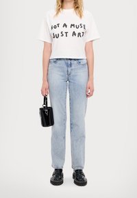 Person klädd i en vit kortärmad T-shirt med texten "INTE EN MUSE, BARA KONST", ljusblå jeans, svarta klumpiga skor, hållande en liten svart handväska vid sidan.