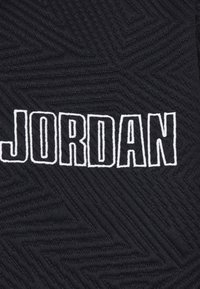 Fekete szövet, amelyen egy struktúrált halszálkamintázat látható, a "JORDAN" szó hangsúlyosan, fehér nagybetűkkel hímezve.