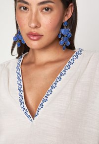 Top bianco con scollatura a V profonda e motivi floreali ricamati in blu. Il modello indossa grandi orecchini a nappina blu. Tessuto dalla consistenza liscia.