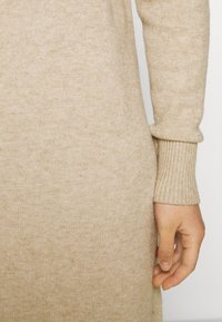 Beżowy sweter dzianinowy, z miękką fakturą i ściągaczami przy mankietach. Zbliżenie dłoni opadającej na materiał, podkreślającej jego gładkie wykończenie.