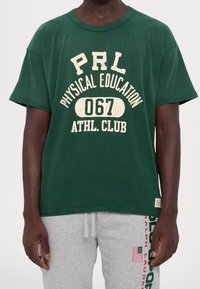 Zelené bavlněné tričko, kulatý výstřih, krátké rukávy, s opotřebovaným bílým textem: "PRL Fyzická výchova 067 Atletický klub." Uvolněný střih.