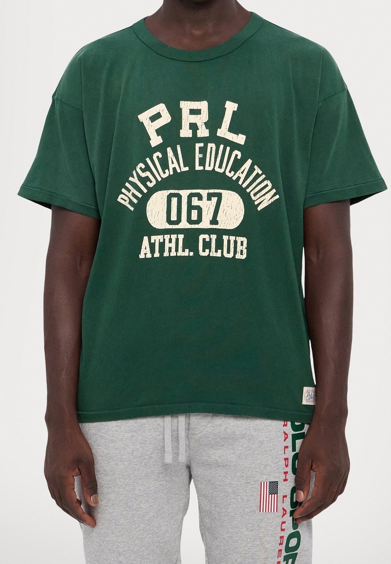 Zelené bavlněné tričko, kulatý výstřih, krátké rukávy, s opotřebovaným bílým textem: "PRL Fyzická výchova 067 Atletický klub." Uvolněný střih.