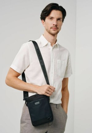 Czarna nylonowa torba na ramię z regulowanym paskiem, gładką teksturą i naszywką z logo z przodu, noszona przez mężczyznę w jasnej koszuli i szarych spodniach.