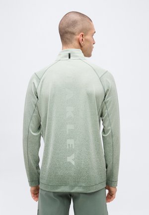 Hombre con la cabeza rapada, vistiendo una camiseta de manga larga verde claro con el texto vertical "OAKLEY" en la espalda y pantalones cortos a juego, de espaldas.