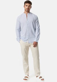 Ung mann med krøllete hår iført en lys blåstripet skjorte med stående krage, beige bukser og hvite sandaler, som står foran en hvit bakgrunn.