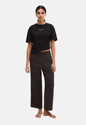 Femme debout pieds nus portant un T-shirt court noir et un pantalon large noir à pois blancs sur un fond uni.