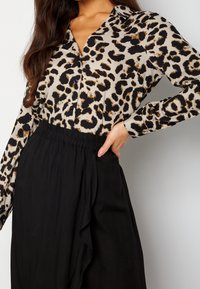 Kvinna iklädd en långärmad blus med leopardmönster, instoppad i lösa svarta byxor med hög midja, poserande med en hand på höften.